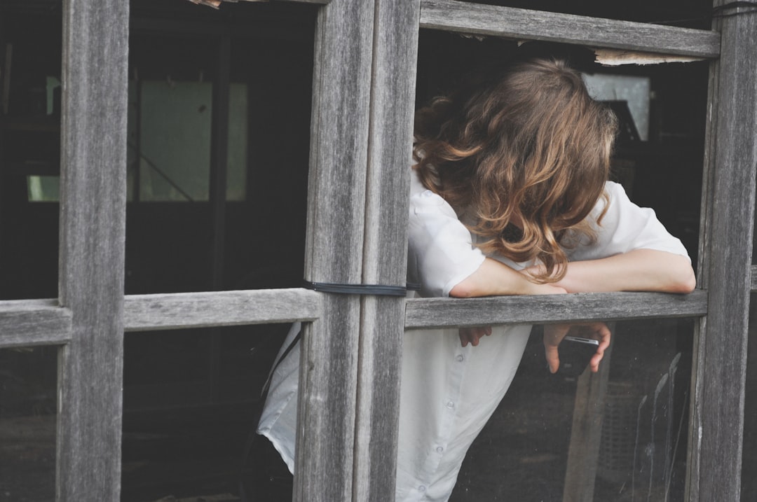 A person stands inside their home looking anxiously out the window, conveying a sense of being stuck and waiting for their house to sell.