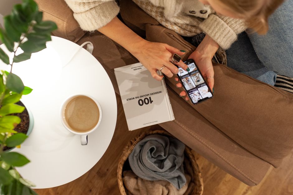 Close-up of a person's hands holding a smartphone, scrolling through listings of beautiful, modern homes for sale on a real estate app.