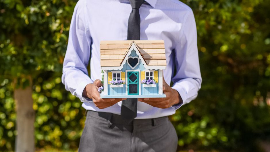 A pair of hands gently cupped around a small model of a house against a clean, bright background, symbolizing the protection of a home's value and a secure real estate transaction.