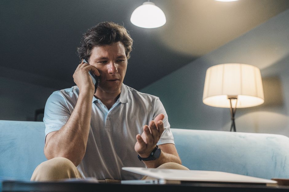 Person sits in a modern living room with a concerned expression while looking at their smartphone, representing the stress...